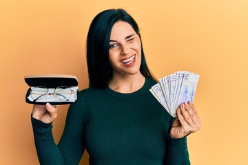 Young caucasian woman holding glasses and swedish krona banknotes winking looking at the camera...