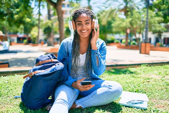 Young african american student woman smiling happy listening to music sitting on the grass at the university campus