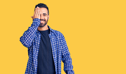 Young hispanic man wearing casual clothes covering one eye with hand, confident smile on face and surprise emotion.