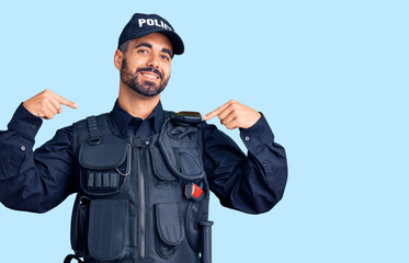 Young hispanic man wearing police uniform looking confident with smile on face, pointing oneself...