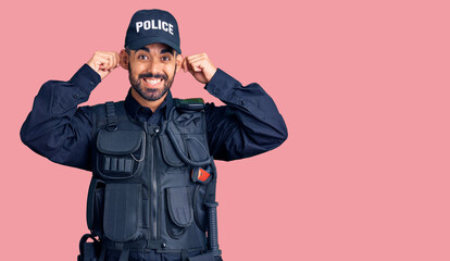 Young hispanic man wearing police uniform smiling pulling ears with fingers, funny gesture. audition problem