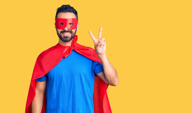 Young hispanic man wearing super hero costume smiling with happy face winking at the camera doing victory sign. number two.