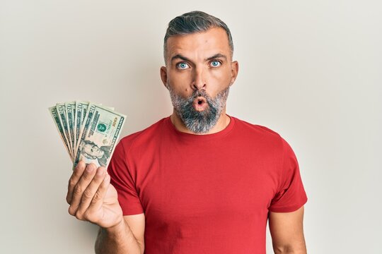 Middle age handsome man holding 20 dollars banknotes scared and amazed with open mouth for surprise, disbelief face