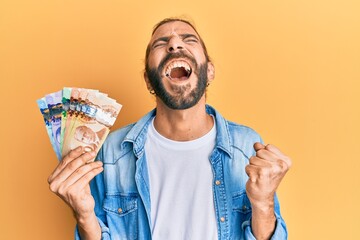 Attractive man with long hair and beard holding canadian dollars screaming proud, celebrating...