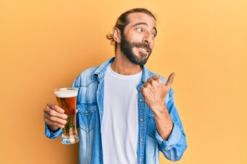 Attractive man with long hair and beard drinking a pint of beer pointing thumb up to the side smiling happy with open mouth