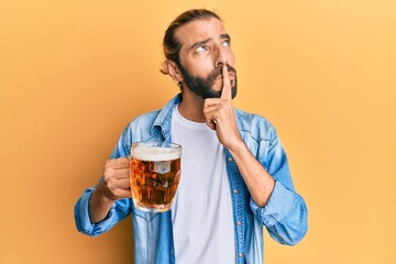 Attractive man with long hair and beard drinking a pint of beer serious face thinking about question with hand on chin, thoughtful about confusing idea