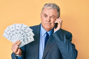 Senior grey-haired man wearing suit holding dollars and using smarpthone winking looking at the...