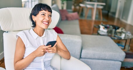 Beautiful brunette woman with short hair at home sitting on the sofa using smartphone