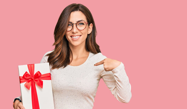 Young brunette woman holding gift pointing finger to one self smiling happy and proud