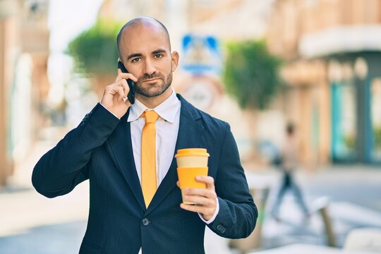 Young hispanic bald businessman with serious expression talking on the smartphone drinking coffee at the city