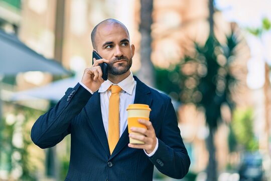 Young hispanic bald businessman with serious expression talking on the smartphone drinking coffee at the city