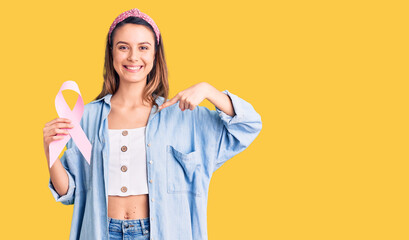 Young beautiful girl holding pink cancer ribbon pointing finger to one self smiling happy and proud