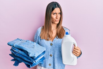 Brunette young woman holding jeans for laundry and detergent bottle skeptic and nervous, frowning...