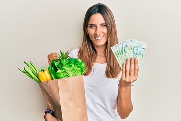Brunette young woman holding groceries and singapore dollars banknotes smiling with a happy and...