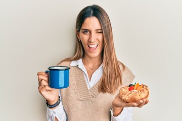 Brunette young woman drinking coffee and eating pastry winking looking at the camera with sexy...