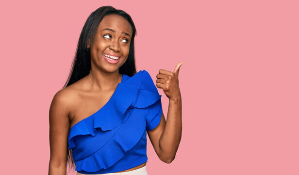 Young black woman wearing casual clothes smiling with happy face looking and pointing to the side with thumb up.
