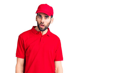 Young handsome man with beard wearing delivery uniform in shock face, looking skeptical and sarcastic, surprised with open mouth