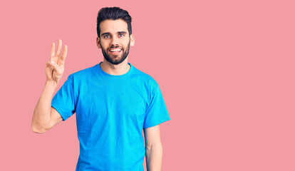 Young handsome man with beard wearing casual t-shirt showing and pointing up with fingers number three while smiling confident and happy.