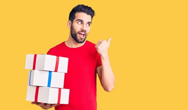 Young handsome man with beard holding gift pointing thumb up to the side smiling happy with open mouth