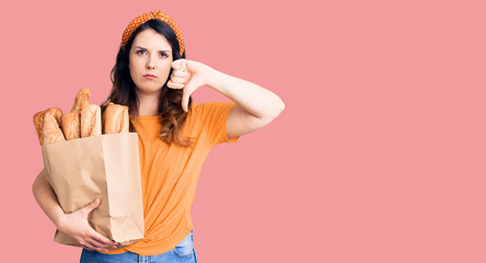 Obraz premium Beautiful young brunette woman holding paper bag with bread with angry face, negative sign showing dislike with thumbs down, rejection concept