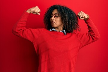 Beautiful african american woman with afro hair wearing sweater and glasses showing arms muscles smiling proud. fitness concept.