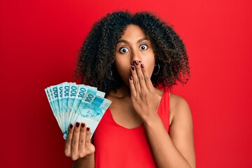 Beautiful african american woman with afro hair holding 100 brazilian real banknotes covering mouth...
