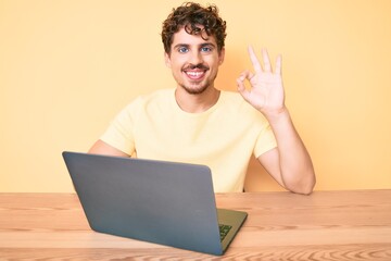 Young caucasian man with curly hair working at the office with laptop doing ok sign with fingers, smiling friendly gesturing excellent symbol