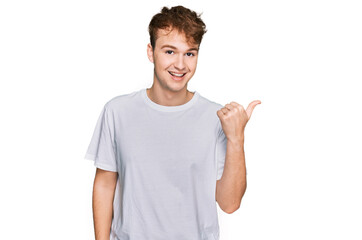 Young caucasian man wearing casual white t shirt smiling with happy face looking and pointing to the side with thumb up.
