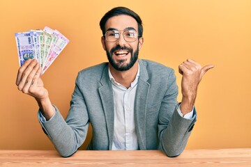 Young hispanic man holding indian rupee banknotes sitting on the table pointing thumb up to the...