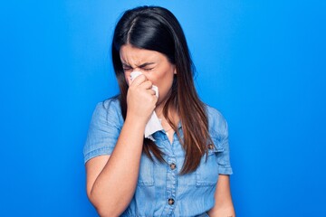 Young beautiful woman sick for flu. Using paper handkerchief on full nose of mucus standing over isolated blue background