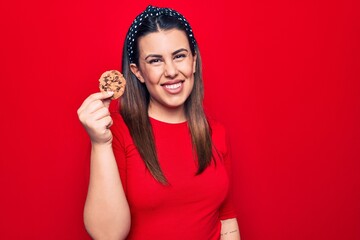 Young beautiful brunette woman holding sweet chocolate cookie over isolated red background looking positive and happy standing and smiling with a confident smile showing teeth