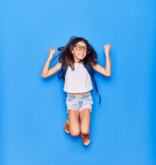 Obraz na płótnie Canvas Adorable hispanic student child girl wearing glasses and backpack smiling happy. Jumping with smile on face over isolated blue background