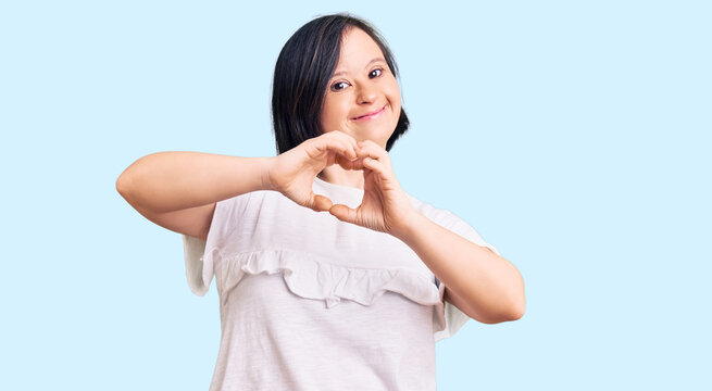 Brunette woman with down syndrome wearing casual white tshirt smiling in love doing heart symbol shape with hands. romantic concept.