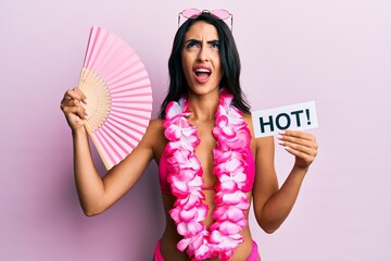 Beautiful hispanic woman wearing bikini and handfan holding hot word angry and mad screaming frustrated and furious, shouting with anger looking up.