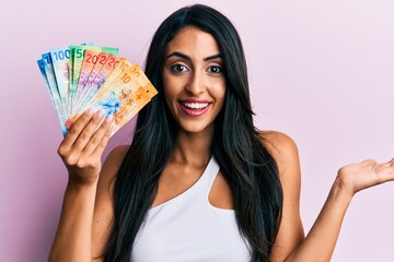 Beautiful hispanic woman holding swiss franc banknotes celebrating achievement with happy smile and winner expression with raised hand