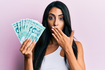 Beautiful hispanic woman holding yuan chinese banknotes covering mouth with hand, shocked and...