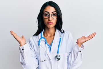 Beautiful hispanic woman wearing doctor uniform and stethoscope clueless and confused with open...
