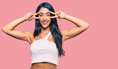 Beautiful hispanic woman wearing casual clothes doing peace symbol with fingers over face, smiling cheerful showing victory