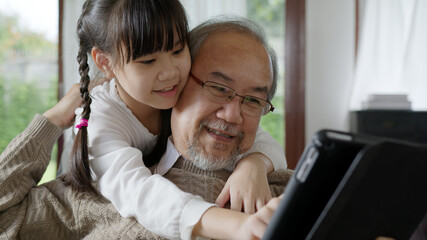 Candid of old senior asian grandparent play and watch with kid grandchildren with technology on computer tablet at home in bonding relationship in family. Young girl hug older man from back. - Powered by Adobe