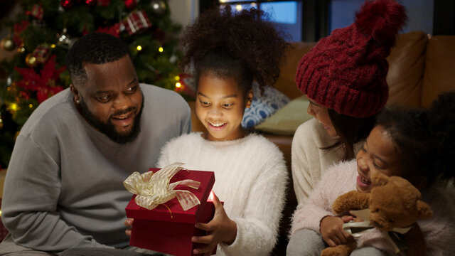 Christmas Day, Happy Black Family At Home. African American Father, Mother And Child Opening Shine Light Xmas Gift Box. Cute Daughter Feeling Excited And Happy With Present.