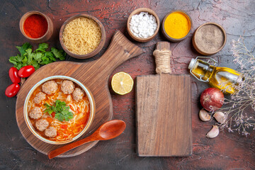 Above view of tomato meatballs soup with noodles in a brown bowl and different spices oil bottle onion garlic and cutting board on dark background