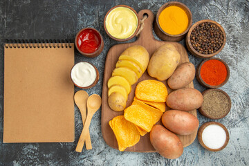 Above view of crispy chips and uncooked potatoes on wooden cutting board different spices and notebook on gray table