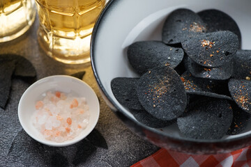 Close-up of a bowl with black potato chips and mugs of beer in the background, selective focus, horizontal shot
