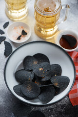 Black potato chips in a bowl with beer and dip, vertical shot on a grey stone background, selective focus