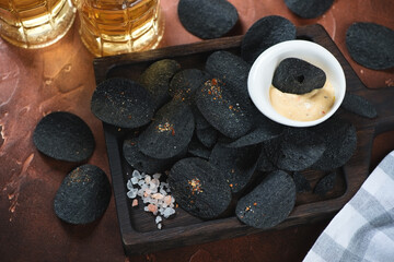 Wooden serving tray with black potato chips and beer mugs in the background, studio shot on a rusty metal surface, elevated view