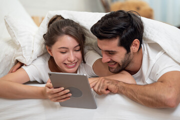Happy young couple looking a tablet together and laughing while lying on the bed