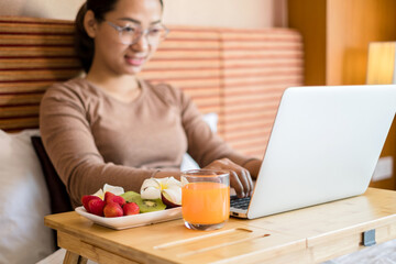 Focus on fruit. young Asian women smiling happily at freelance work, working on a notebook while relaxing in bed with snacks and fruit in a hotel room. Vacation and relaxation