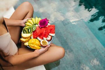 Young unrecognizable woman by the pool with a plate of tropical fruits: watermelon, pineapple,...