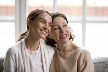 Close up head shot smiling dreamy grownup daughter and mother dreaming together, hugging, sitting on cozy couch at home, two generations visualizing future, enjoying leisure time, relaxing