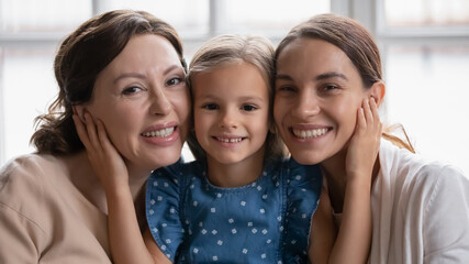 Head shot portrait smiling three generations of women posing for family photo together, happy little girl with mature grandmother and young mother looking at camera, touching cheeks, enjoying weekend